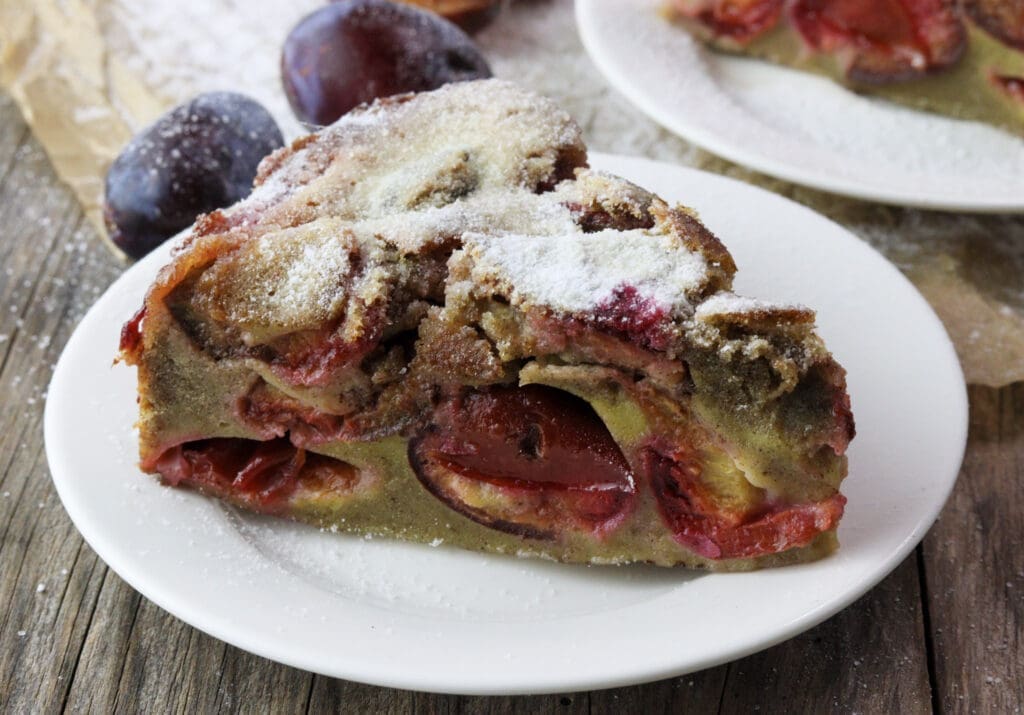 A juicy slice of buckwheat plum clafoutis with plums in the background.